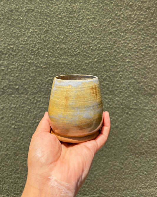 A hand holding a small handmade ceramic cup with a textured, speckled glaze in earthy yellow, brown, and gray tones, set against a matching textured olive-green wall.