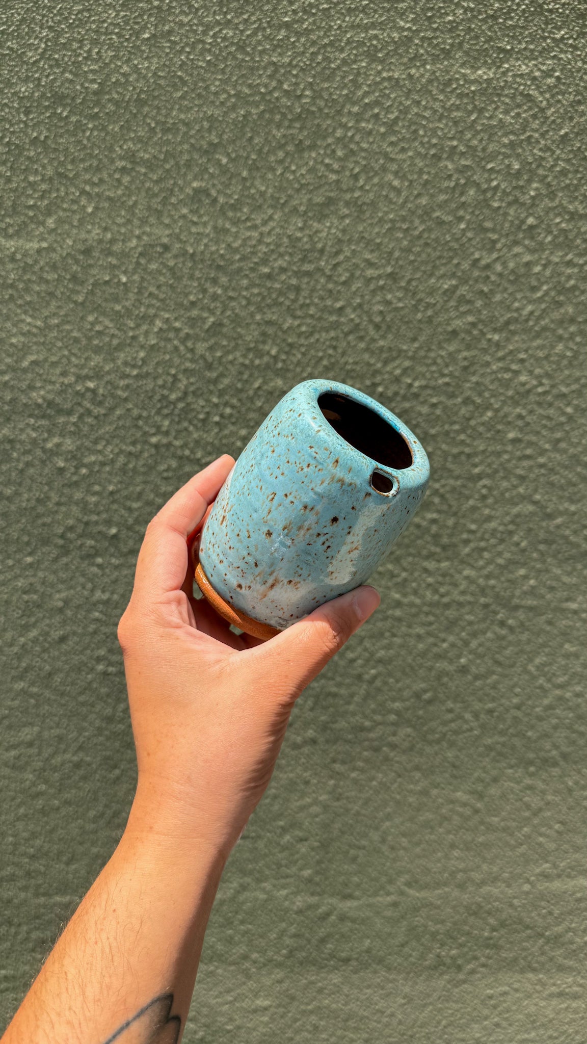 A hand holding a small cylindrical ceramic cup with a speckled light blue glaze and a small brown base, set against a textured olive green wall.