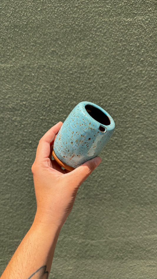 A hand holding a small cylindrical ceramic cup with a speckled light blue glaze and a small brown base, set against a textured olive green wall.