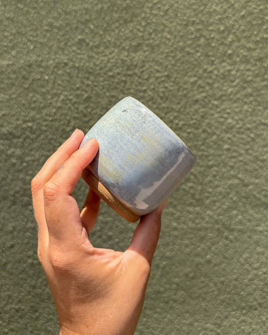 A hand holding a ceramic cup with a light blue and white marbled glaze, set against a textured olive green wall.